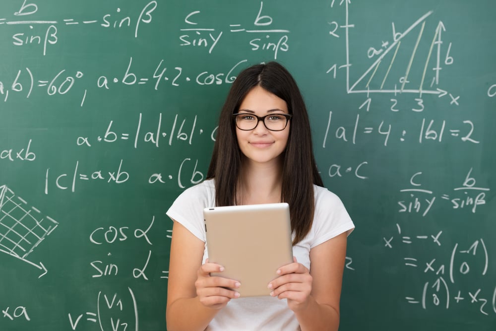 Clever attractive young female college student wearing glasses and holding a tablet computer in her hands standing in front of a chalkboard covered in equations in maths class smiling at the camera-1