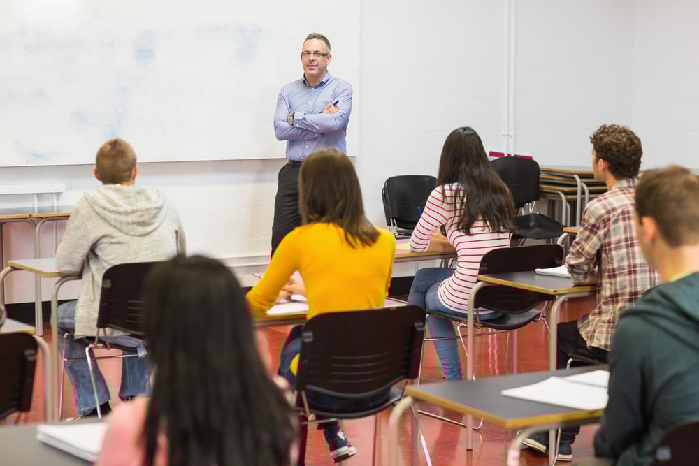 Rear view of students attentively listening to male teacher in the classroom